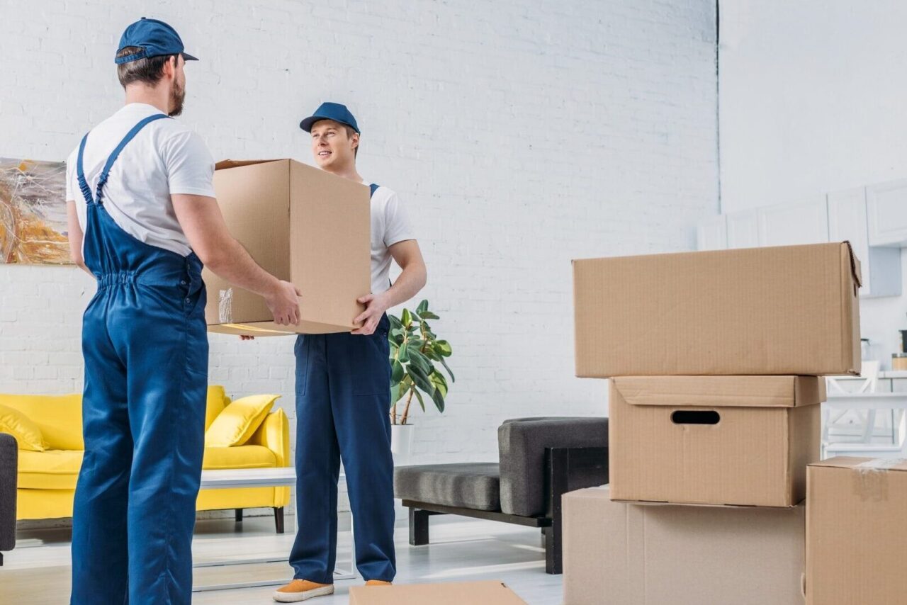 two movers in uniform transporting cardboard box in living room