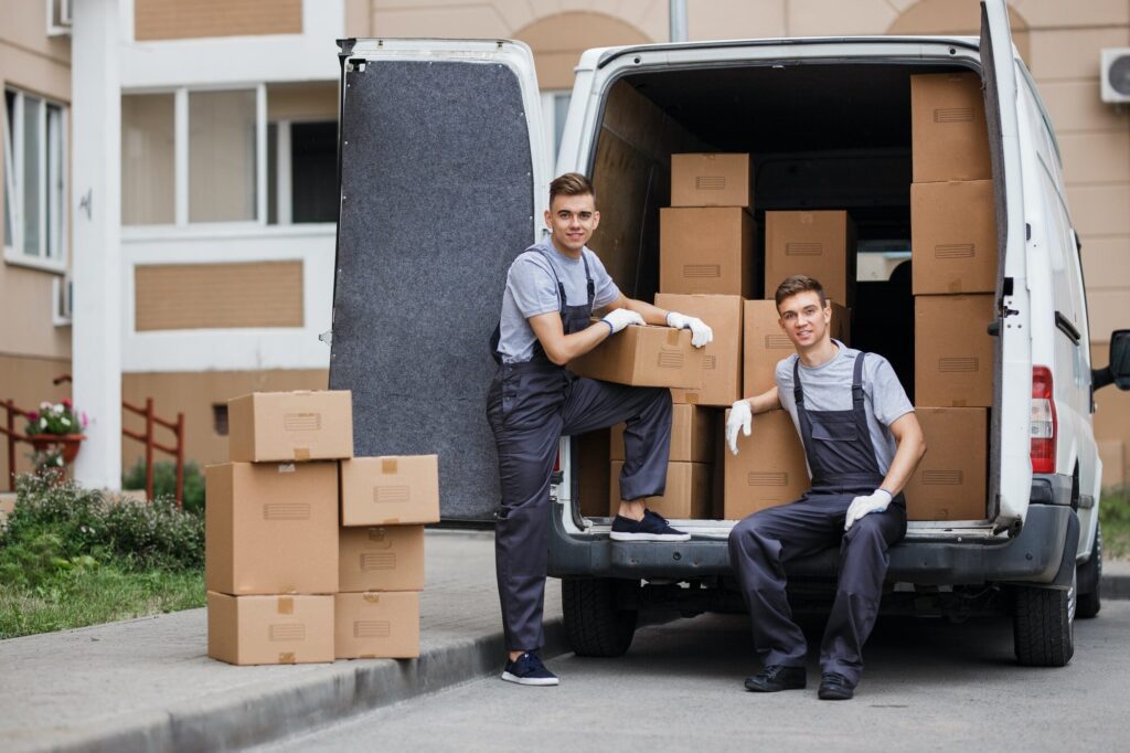 Two young handsome smiling movers wearing uniforms are unloading the van full of boxes. House move