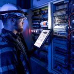 Electrician in overalls, focused on work in switchboard with fuses, using tablet.