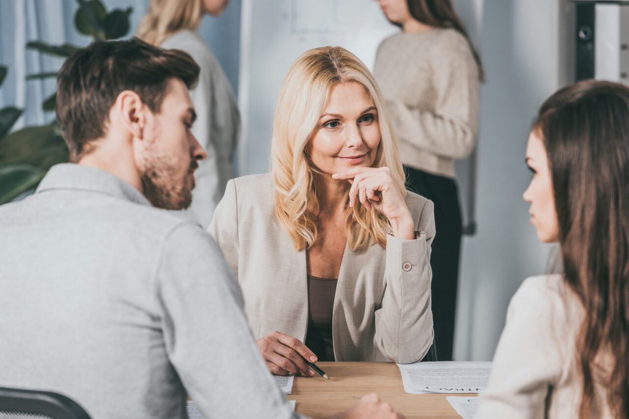 beautiful smiling business mentor sitting with hand on chin and looking at young colleagues in