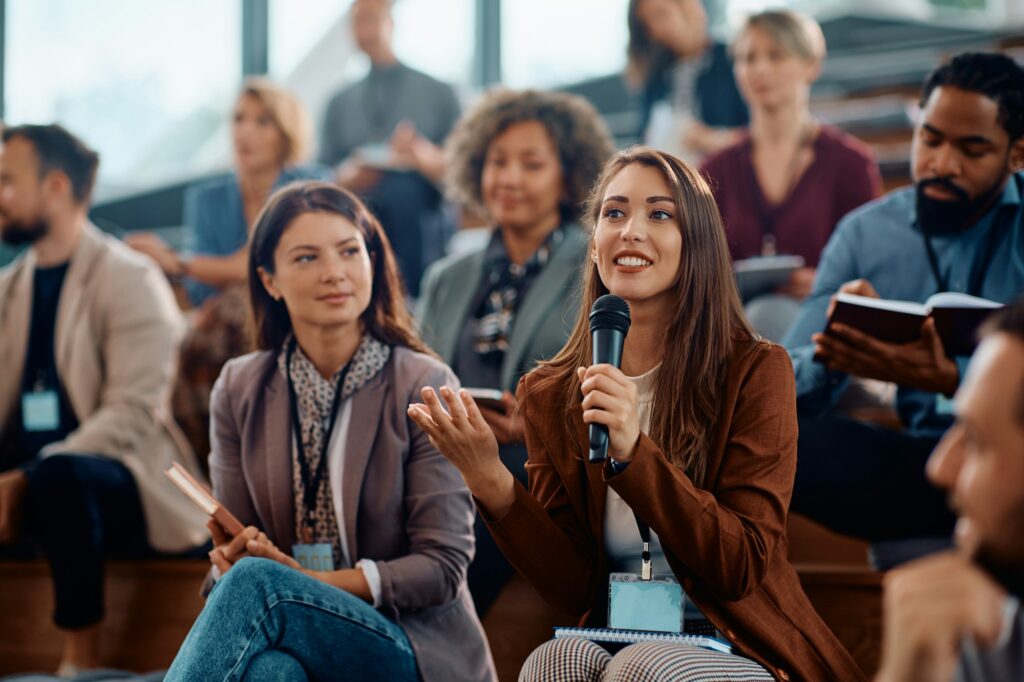 Young happy businesswoman talking on microphone while attending education event in conference hall.