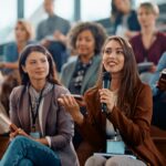 Young happy businesswoman talking on microphone while attending education event in conference hall.