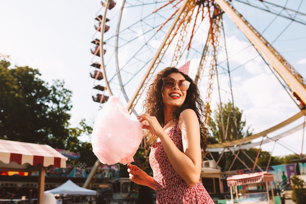 Happy girl in sunglasses and birthday cap eating cotton candy in amusement park over ferris wheel