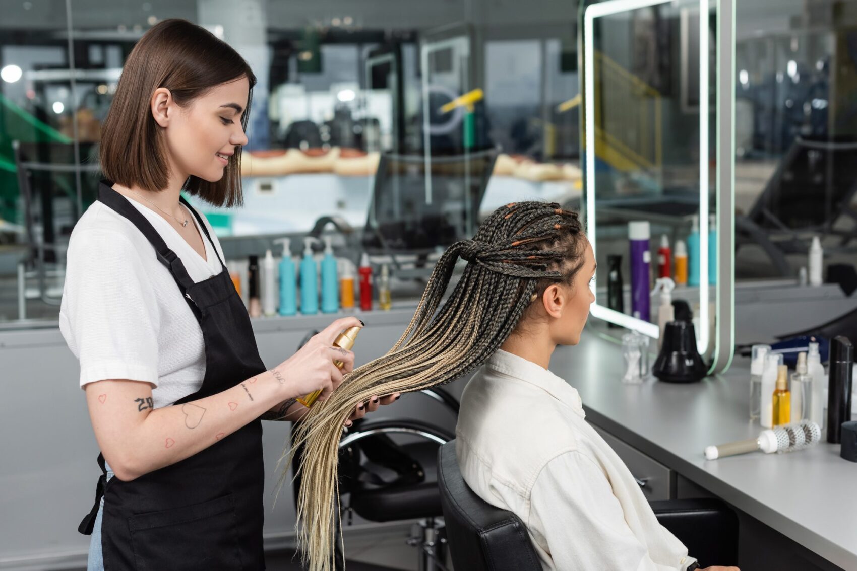 hair salon, tattooed beauty worker in apron applying hair product on braids of woman in salon, spray